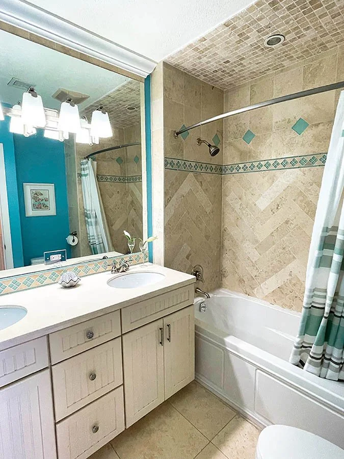 Bathroom with a double sink vanity, large mirror, and a bathtub with a shower curtain. The shower has beige tile walls with decorative blue accents.