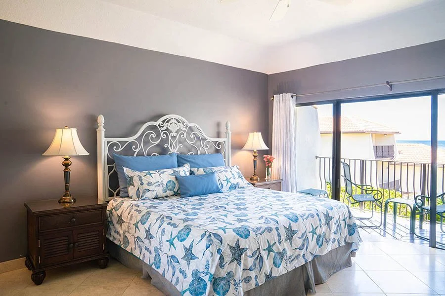 Bedroom with a white ornate bed frame, blue and white starfish-themed bedding, two bedside tables with lamps, and sliding glass doors leading to a balcony with chairs and a view of neighboring houses and the ocean.