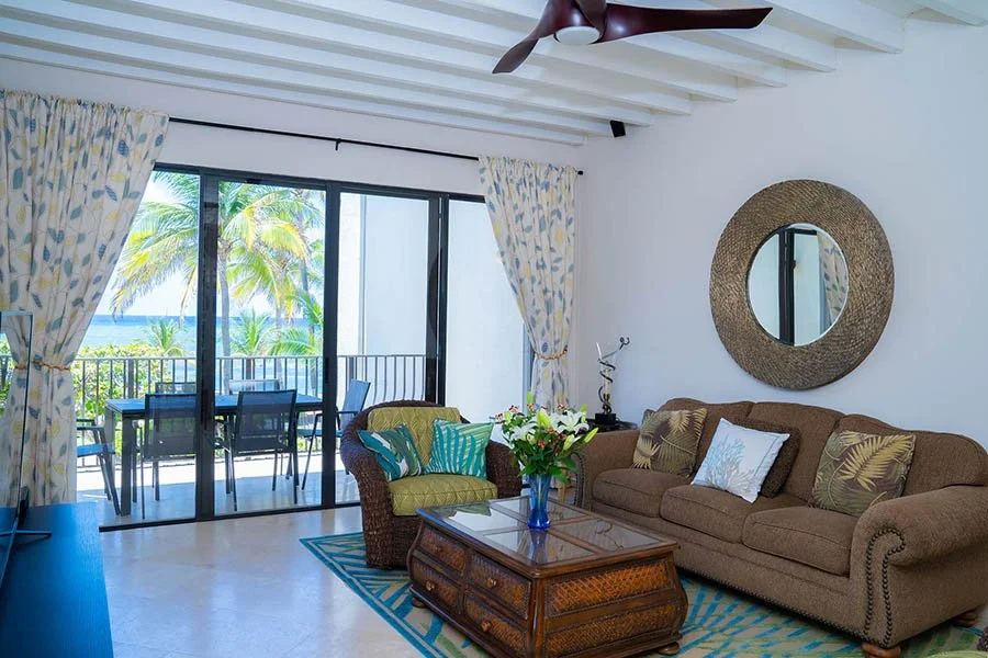 Living room with sliding glass doors leading to a balcony with outdoor seating, tropical view, wicker and fabric sofa, armchair, wooden coffee table, decorative mirror, tropical-themed cushions, flowers in a vase, ceiling fan, and tropical curtains.