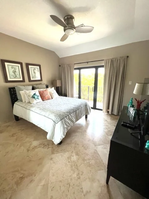 Bedroom with a large bed, framed animal artwork on the wall, a ceiling fan, sliding glass door with curtains leading to a balcony, and a black dresser with decorative items.