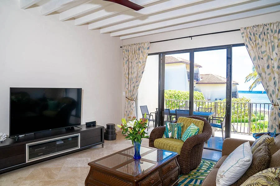 Living room with sliding glass doors opening to balcony with outdoor seating, watching ocean view, with sofa, armchair, coffee table with flowers, television, and curtains.