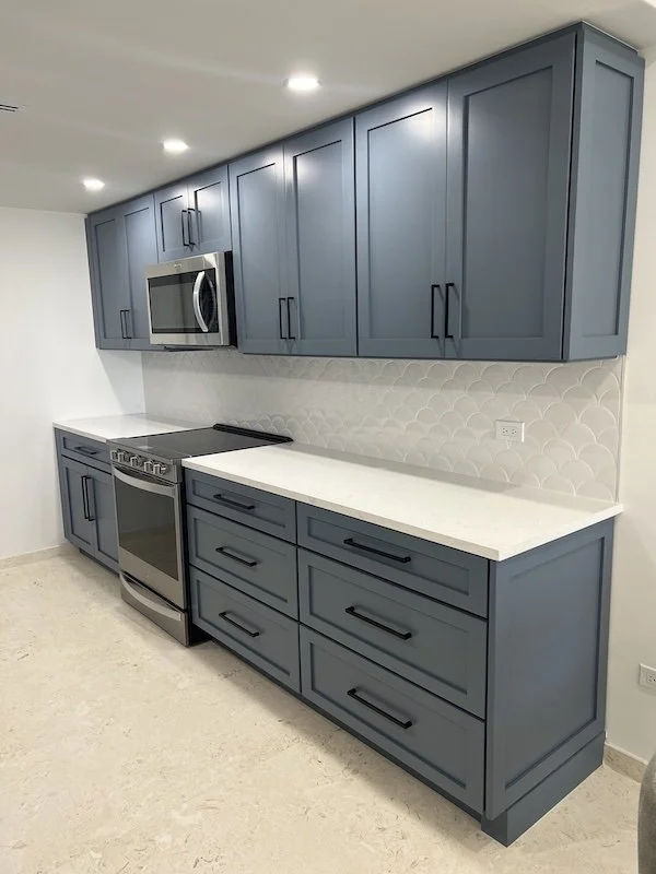 Modern kitchen with blue cabinets, white countertop, stainless steel microwave and oven, white tile backsplash, and beige floor.