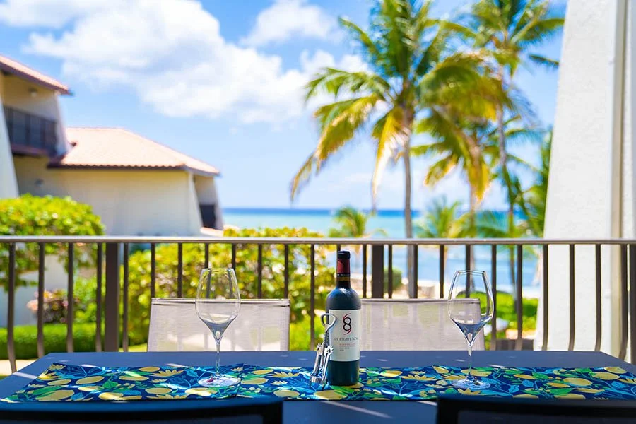 Outdoor balcony table set for two with wine bottle and glasses, overlooking tropical palm trees and ocean view.