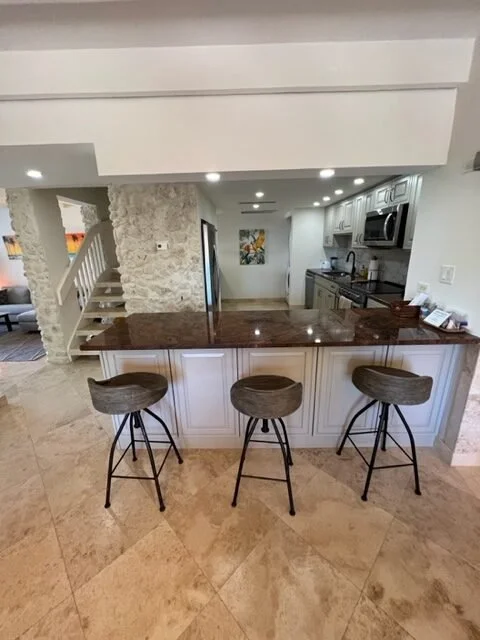 Modern kitchen with granite island, three bar stools, white cabinetry, stainless steel appliances, and recessed ceiling lights.