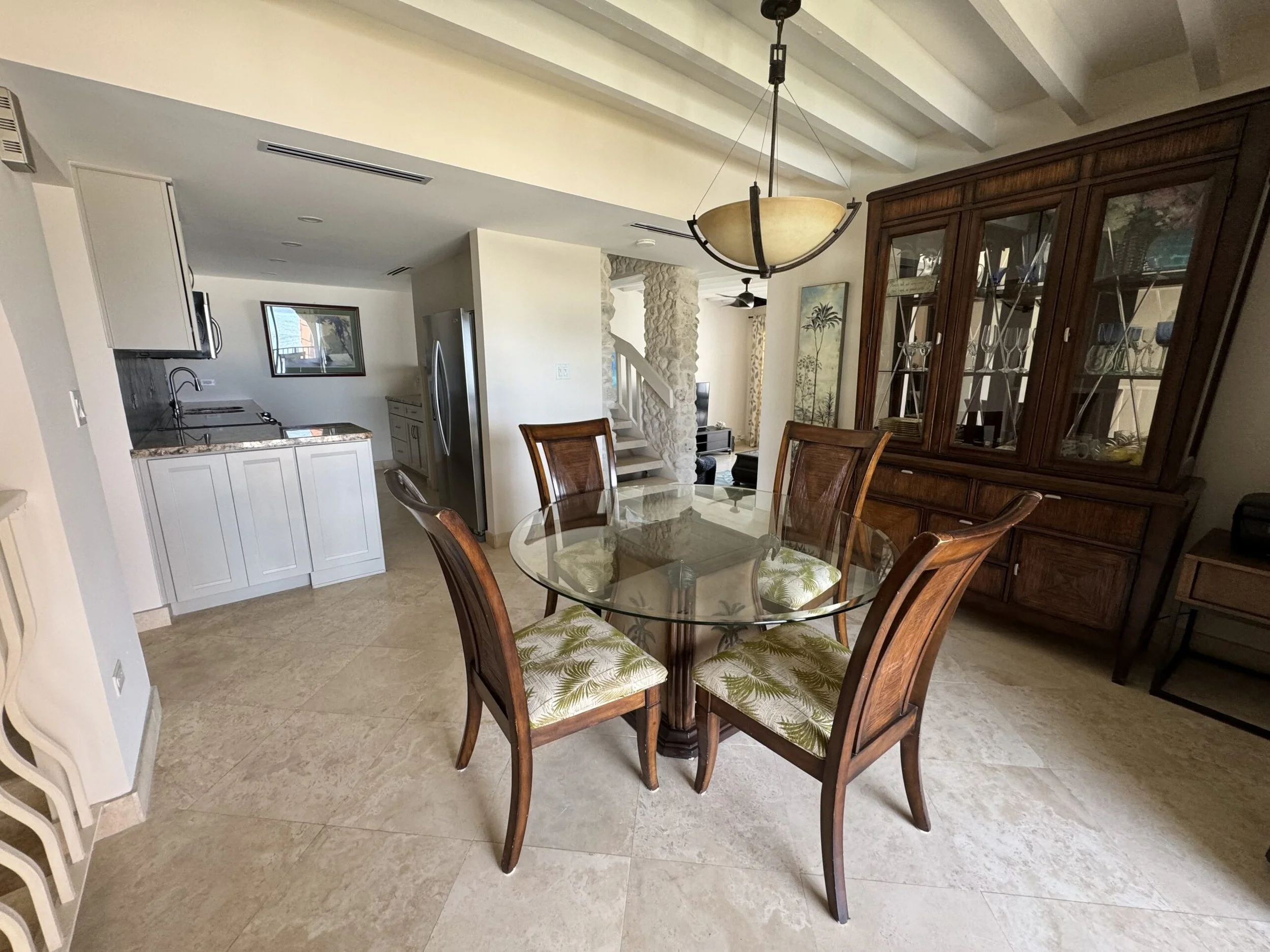 A dining area with a round glass table and four wooden chairs with patterned cushions. A large wooden china cabinet with glass doors displays glassware. Overhead, a modern bowl-shaped chandelier hangs. The background includes part of the kitchen with