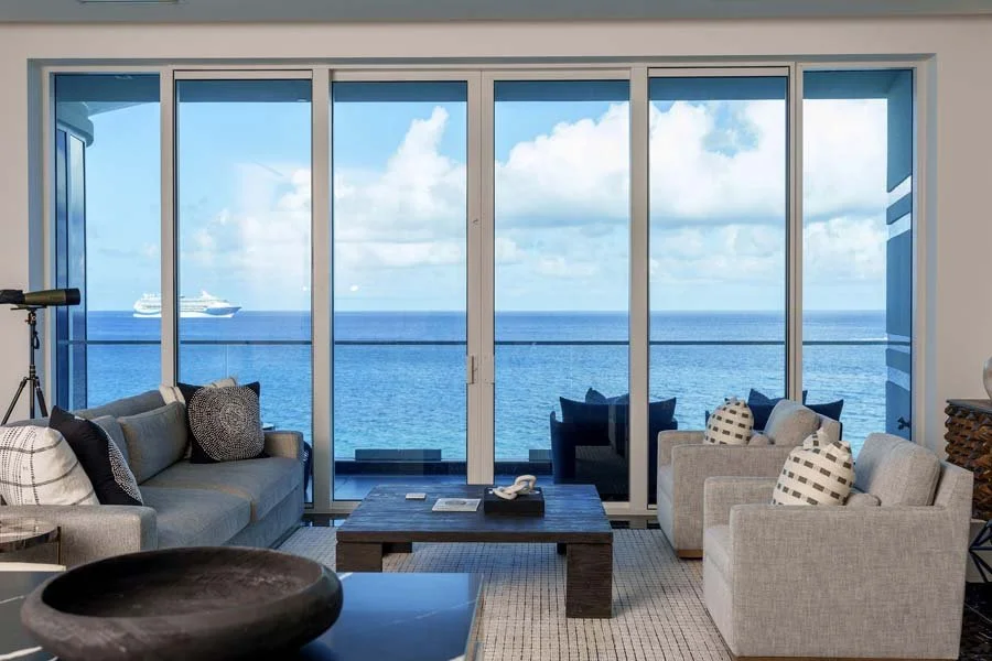 Living room with large glass sliding doors overlooking the ocean and a cruise ship in the distance. Contains sofas, armchairs, a coffee table, and a telescope.