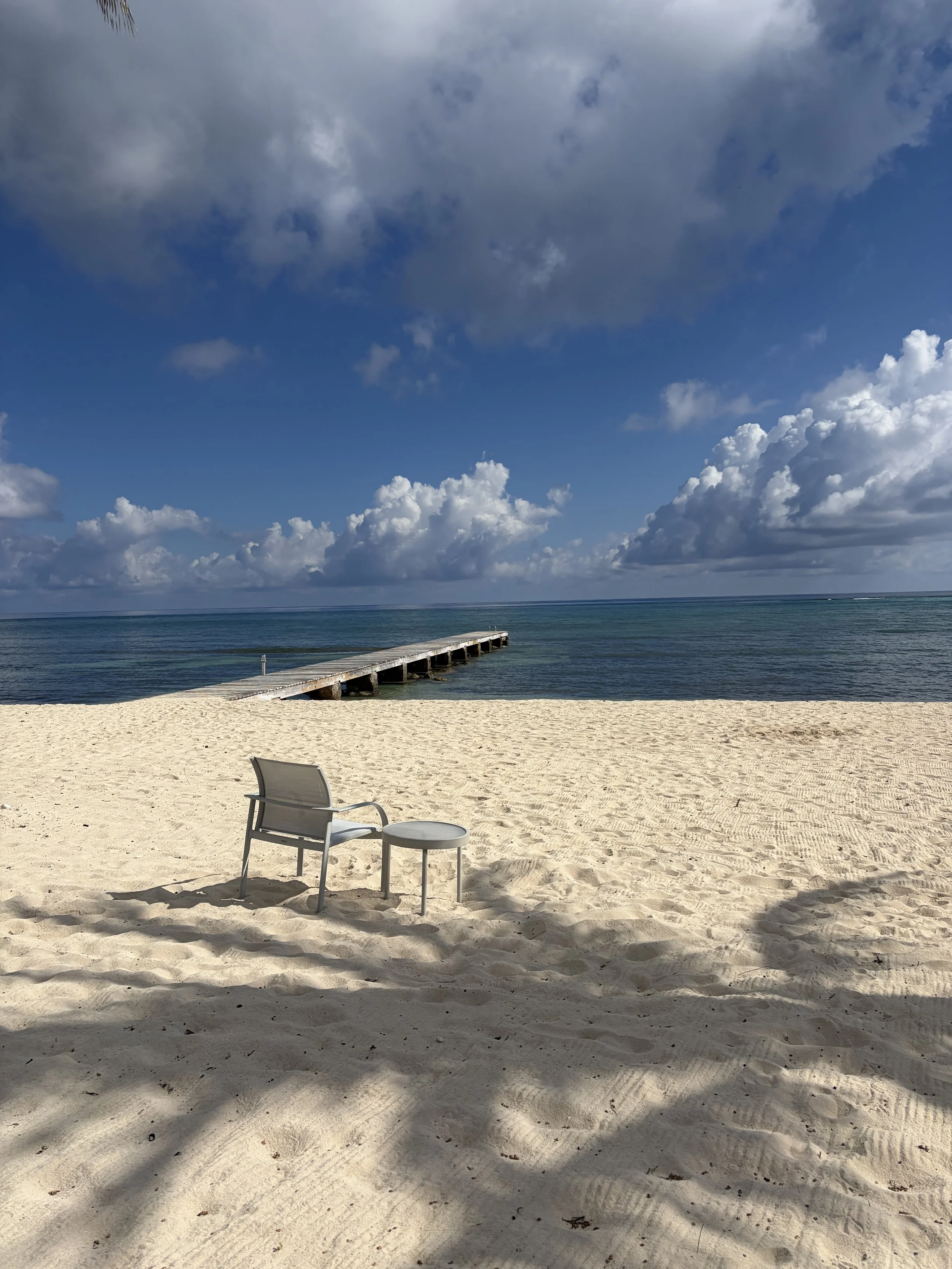 A sandy beach with a single chair and small table in the foreground, a wooden pier extending into the ocean, and a partly cloudy sky overhead.