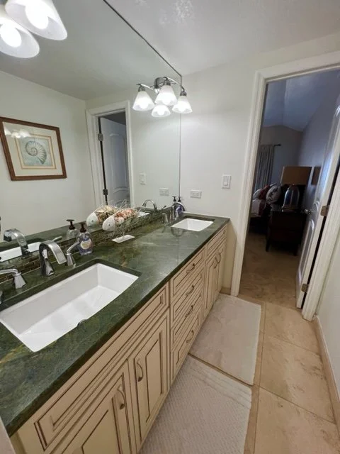 Bathroom vanity with a green countertop, two sinks, and a mirror with a framed picture on the wall, visible through an open door.