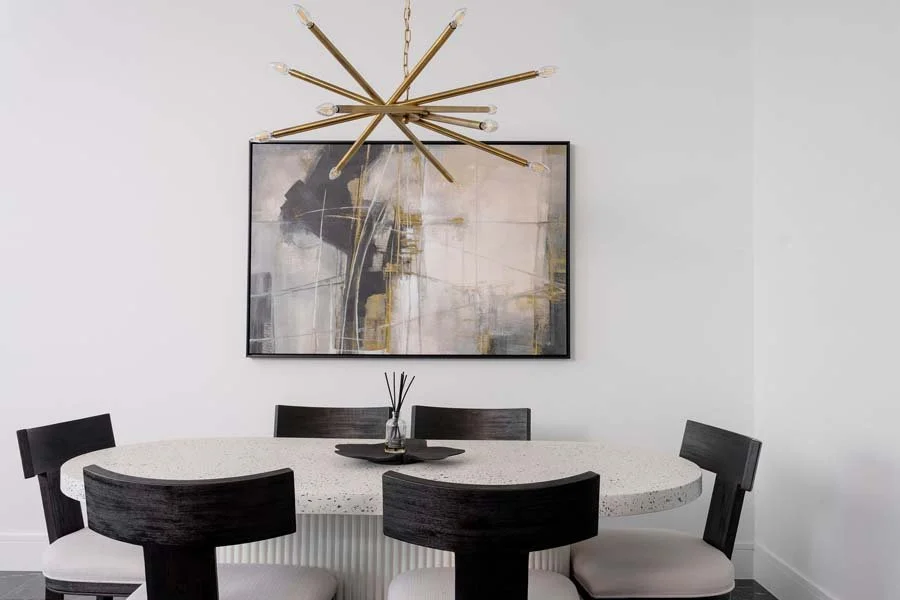 Modern dining room with a white oval table, six black chairs with white cushions, abstract painting on a white wall, and a gold chandelier overhead.