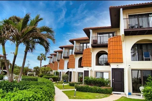 Beachside hotel or apartment complex with white walls, orange tiled awnings, and balconies, surrounded by palm trees and landscaped greenery under a blue sky.