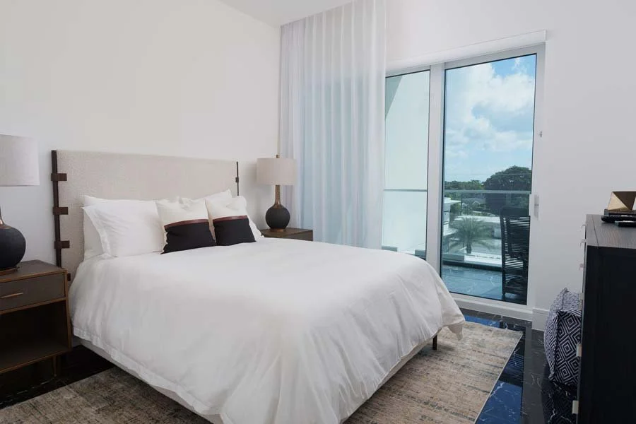 A modern bedroom with a balcony, white bed with pillows, two bedside tables with lamps, and a view of trees and sky through the glass sliding door.