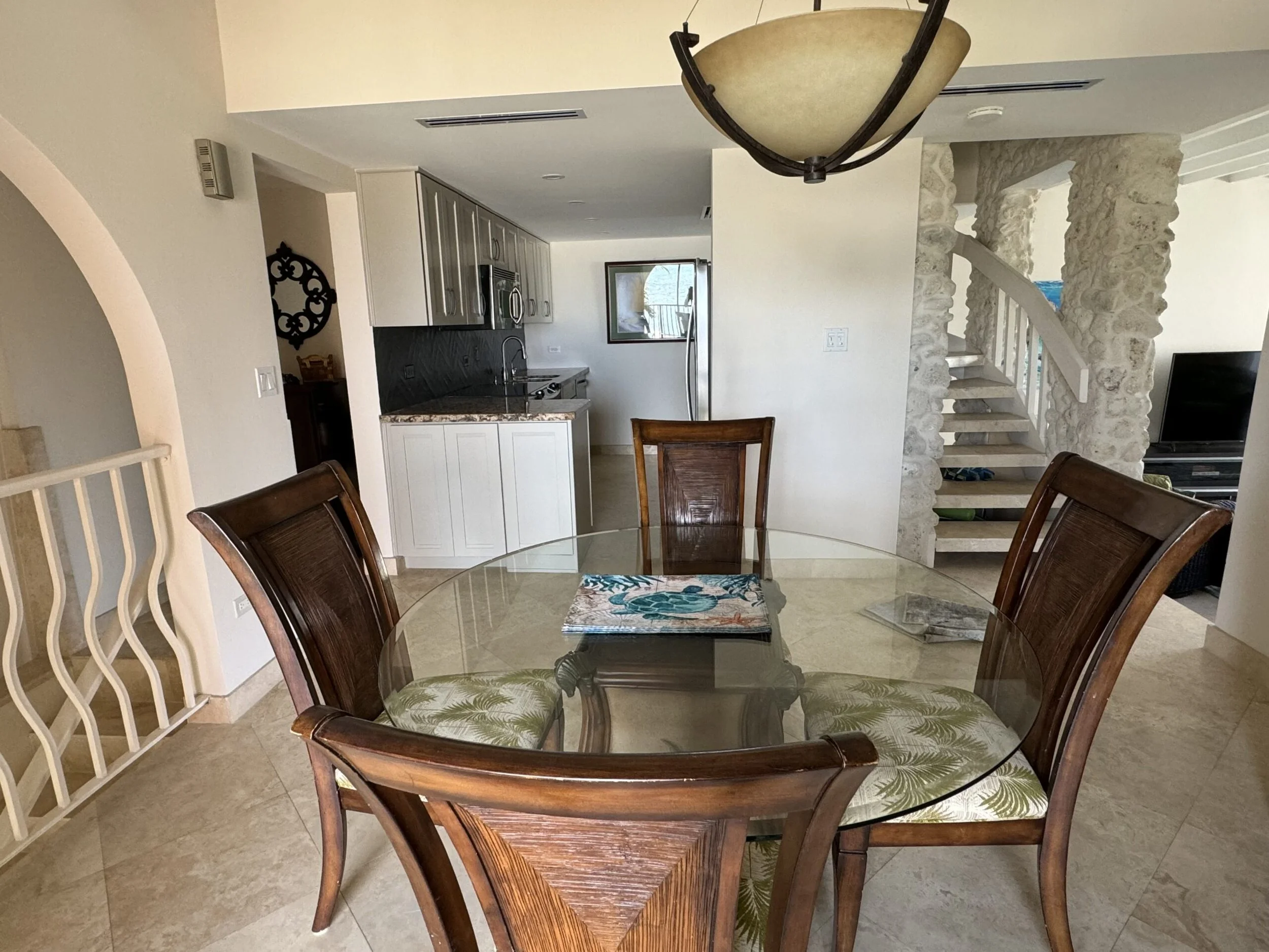 A dining area with a glass-top wooden table and six matching wooden chairs with green leaf-patterned cushions. There is a decorative rug underneath the table. The background reveals a kitchen with grey cabinets, a marble countertop, and a window with