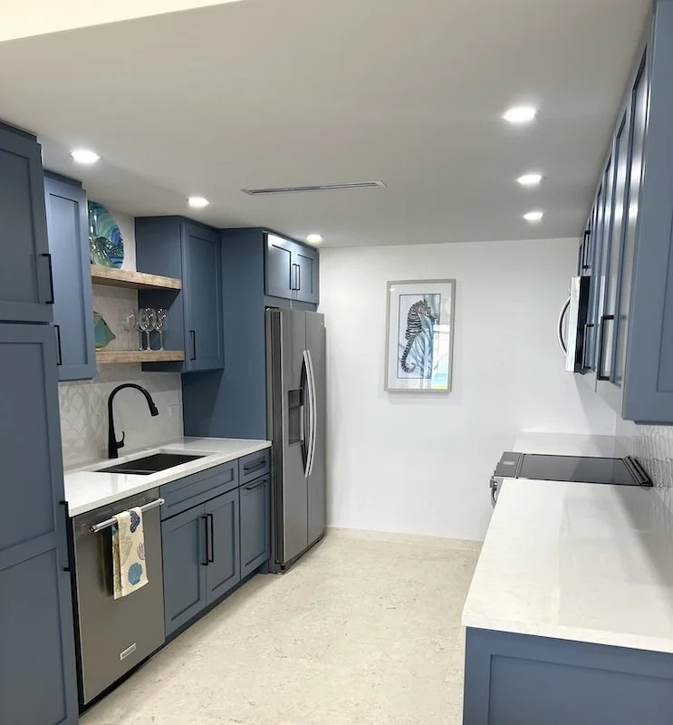 Modern kitchen with blue cabinets, a stainless steel refrigerator, a white countertop, a black sink with a black faucet, a framed zebra artwork on the wall, and a small open shelf with glassware.