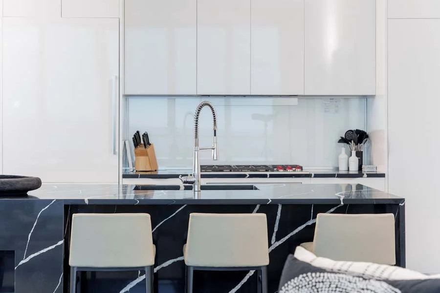 Modern kitchen with white cabinetry, black marble island with white veining, and a sink with a flexible faucet. There are three beige chairs at the island, and a knife block, utensils, and bottles on the counter.