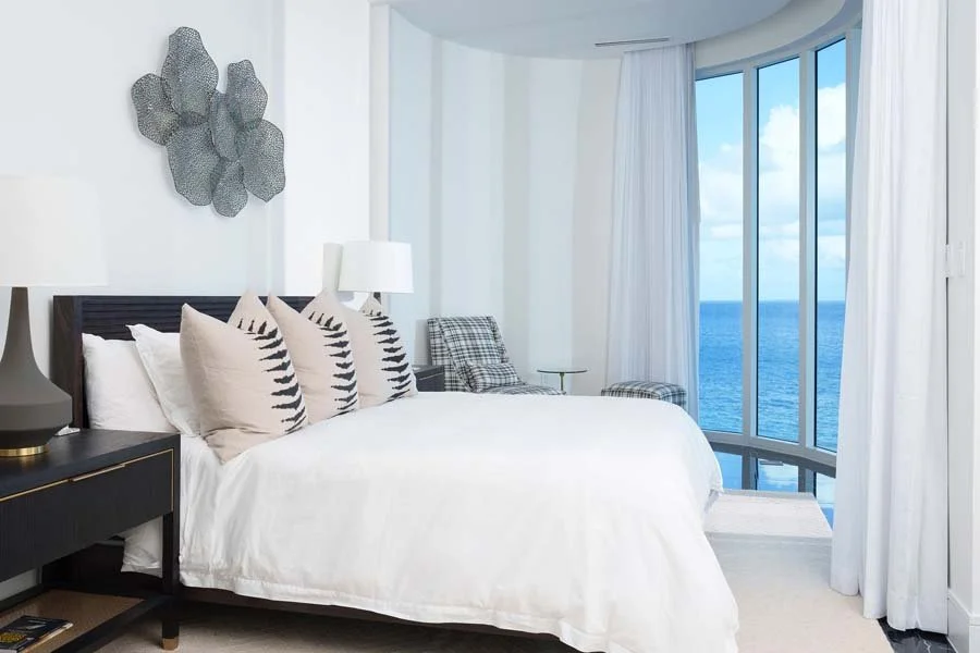 A modern hotel bedroom with a large window view of the ocean, white walls, a bed with white linens and decorative pillows, a bedside table with a lamp, and a seating area with chairs and a small table.