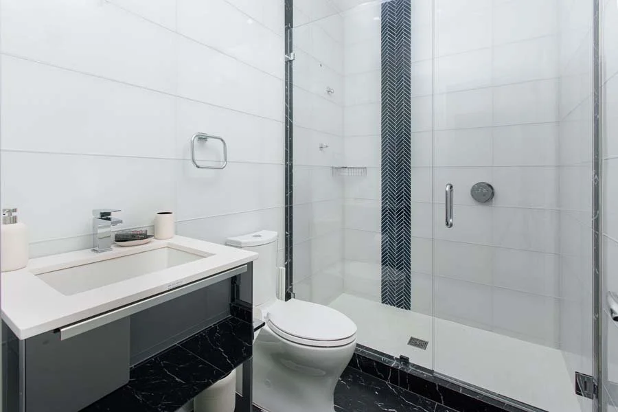 Modern bathroom with white tiled walls, black marble floors, glass shower enclosure, white toilet, and a vanity with a rectangular sink and black decorative panel.