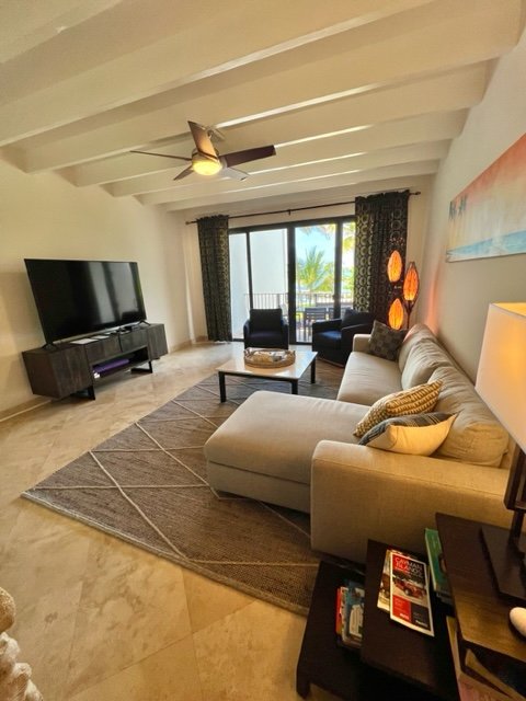 Living room with beige sectional sofa, black armchairs, and a large flatscreen TV on a stand. There is a sliding glass door leading to a balcony with palm trees visible outside. The room has a ceiling fan, a patterned rug, and decorative lamps.