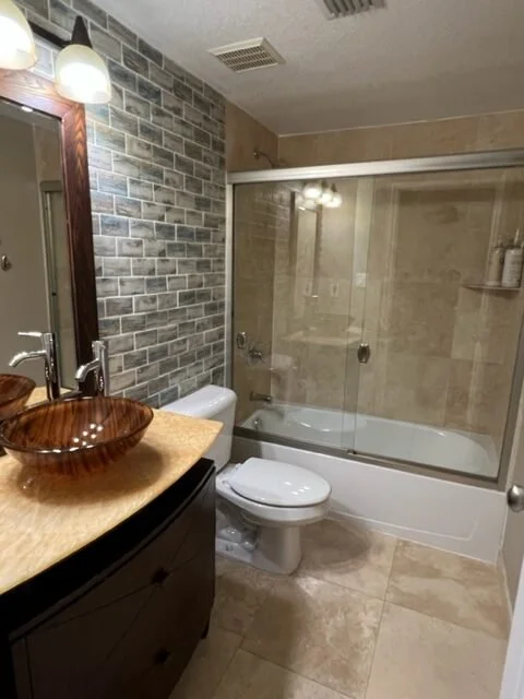 Bathroom with brick accent wall, wooden vessel sink on granite countertop, mirror, toilet, glass-enclosed bathtub, and beige tile flooring and walls.