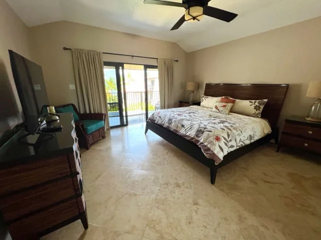 A bedroom with a large bed, nightstands with lamps, a ceiling fan, a chair near sliding glass doors, and a view of a balcony with palm trees outside.