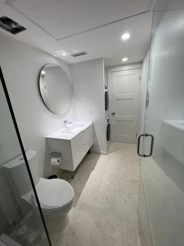 Modern white bathroom with toilet, floating vanity with sink, round mirror, laundry area with stacked washer and dryer, and a glass shower door.