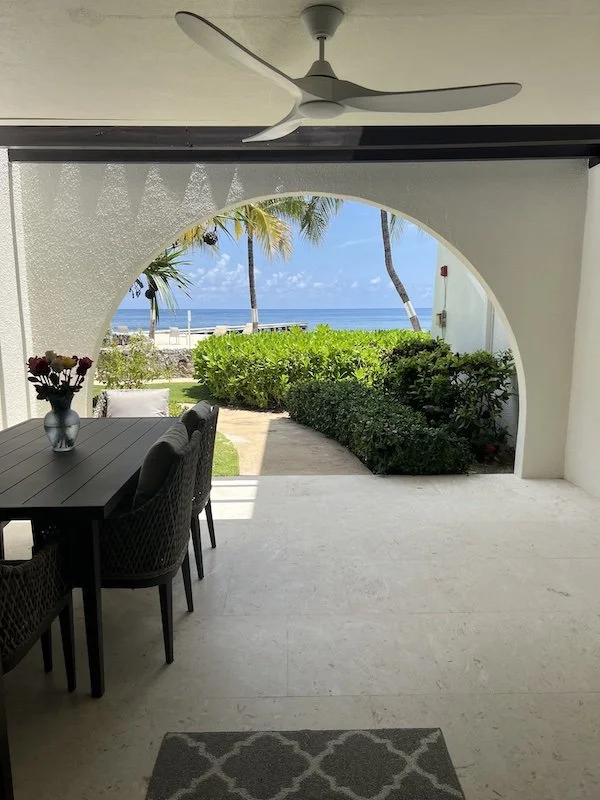 View of a beach through an arched opening from a shaded patio with a dining table and chairs, ceiling fan, and nearby bushes and palm trees.