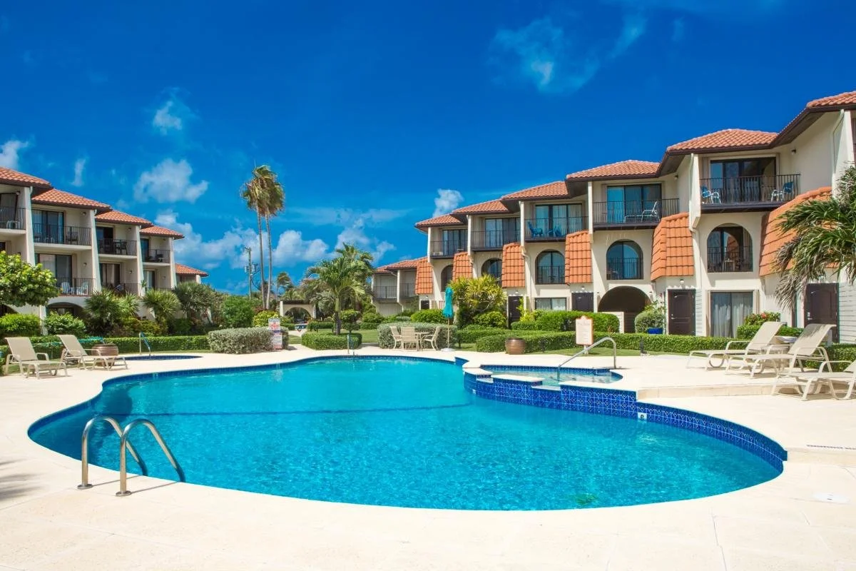 A resort-style outdoor swimming pool with lounge chairs, surrounded by apartment buildings with balcony views, under a bright blue sky with a few clouds and palm trees.