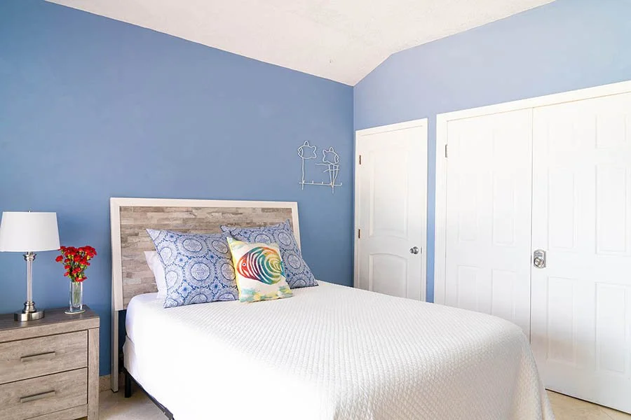 A bedroom with a blue wall, whitebed with a wooden headboard, three pillows (two blue patterned and one with colorful abstract design), a nightstand with a lamp and small red flowers, and two white closet doors.
