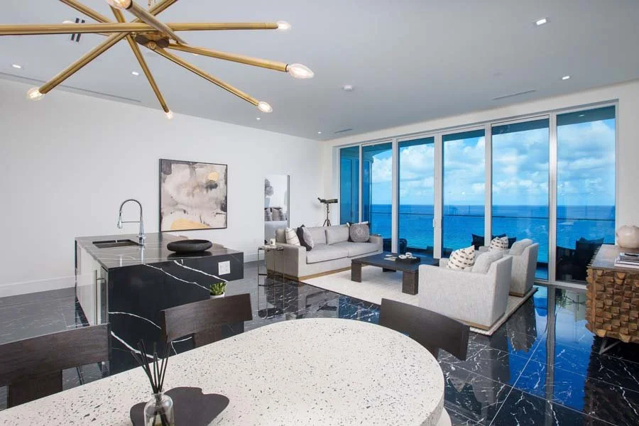 Modern living room with panoramic ocean view, white sofas, black coffee table, abstract wall art, and a glass sliding door leading to a balcony.