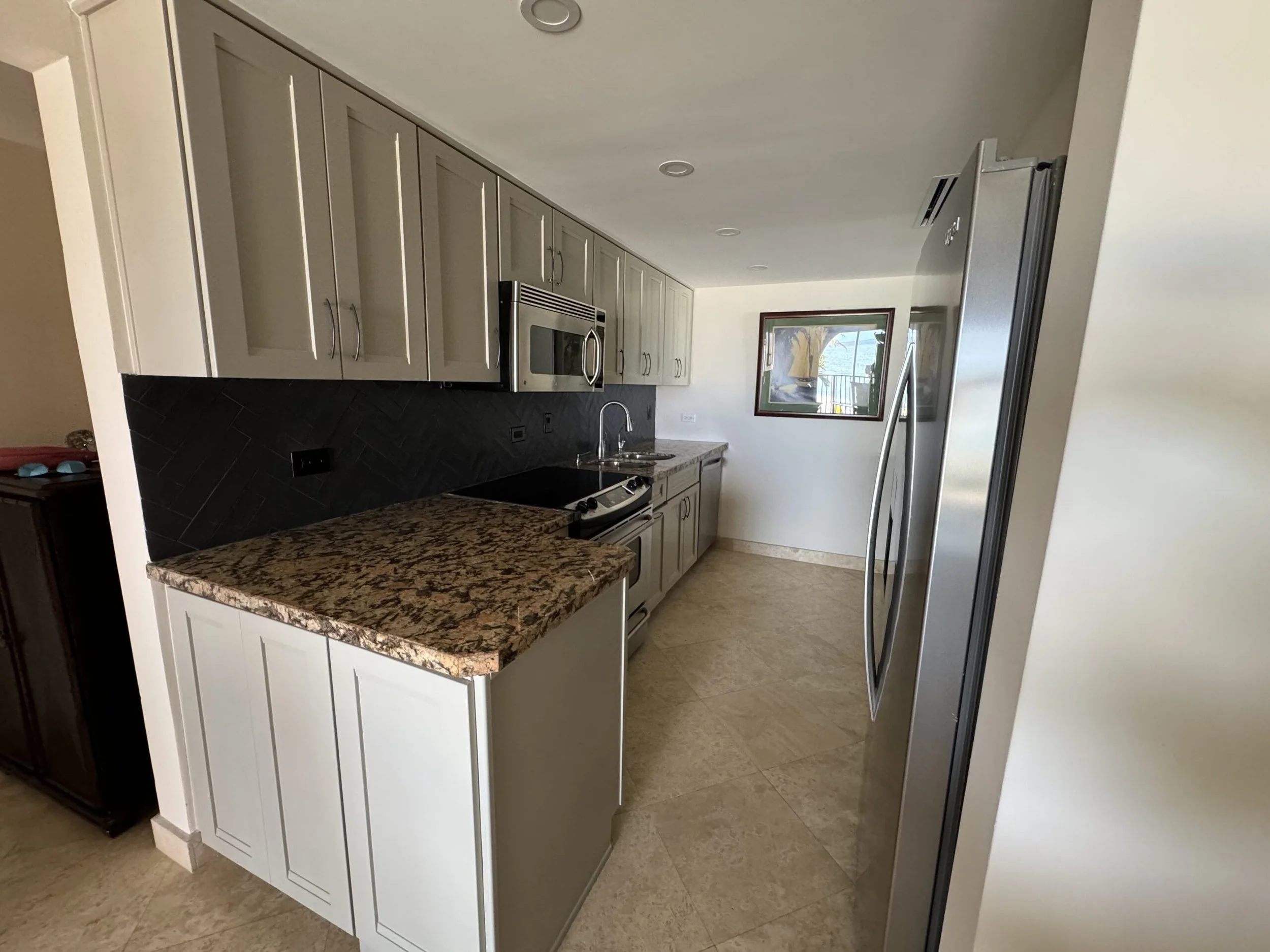 A modern kitchen with beige cabinets, a granite countertop, and stainless steel appliances including a microwave and refrigerator. There is a window with a view outside.