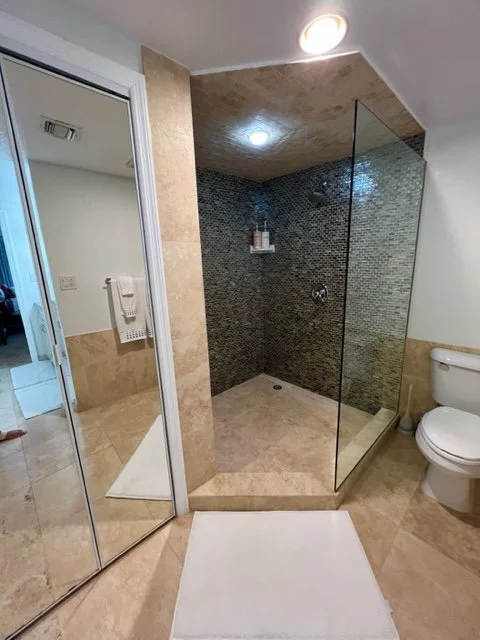A bathroom with a walk-in shower, glass partition, beige tile flooring, and a toilet. There's a mirror on the left wall and a towel rack with a towel visible.