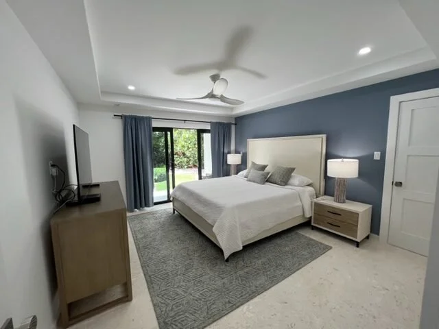 Modern bedroom with a large bed, two nightstands with lamps, a dresser with a TV, sliding glass door with blue curtains, and a ceiling fan.
