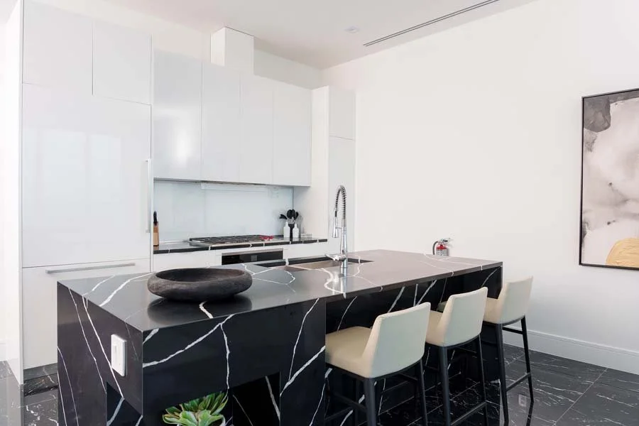 Modern kitchen with black marble island, white cabinets, beige chairs, and minimalist decor.