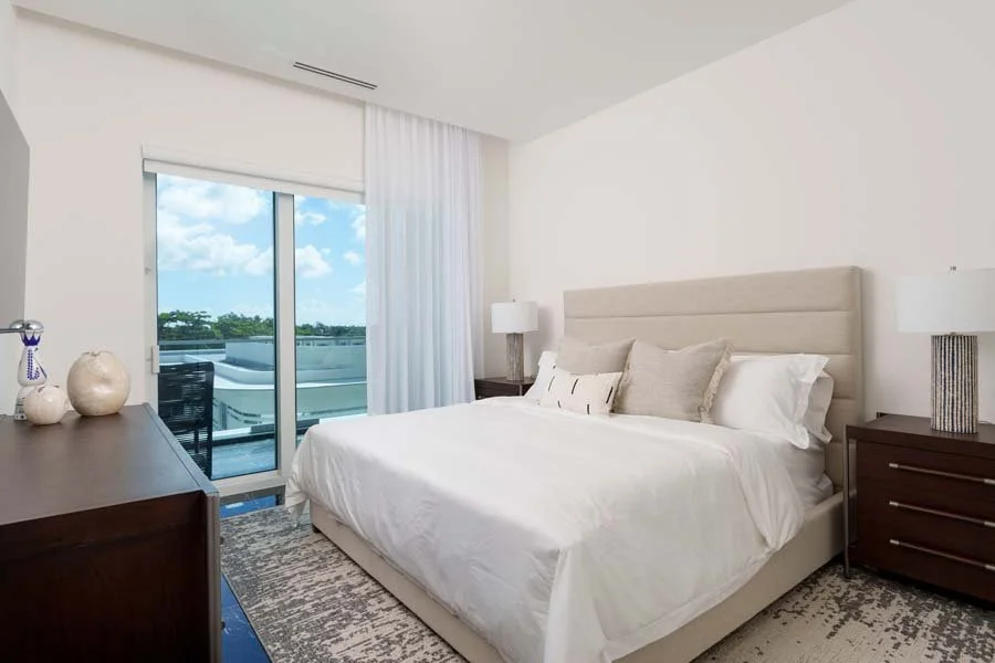 Modern bedroom with a large bed, beige headboard, white bedding, and decorative pillows. There are two matching nightstands with lamps, a dresser with decorative vases, and a sliding glass door with curtains opening to a balcony with a view of trees 