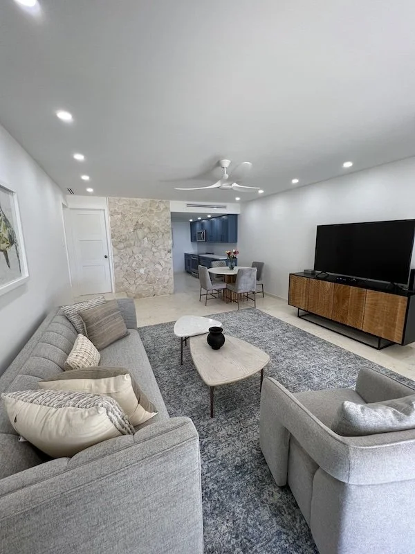 Living room with gray sofa and armchair, coffee table, large flat-screen TV on a wooden stand, art on the wall, and a dining area with a round table and four chairs, opening to a kitchen.