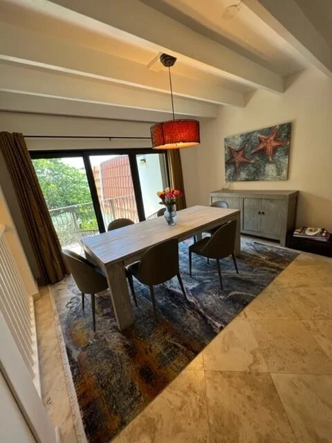 Dining room with a rectangular wooden table, four chairs, a colorful rug, a sliding glass door to a balcony, a hanging red lampshade, a sideboard with a starfish painting, and a vase of flowers on the table.