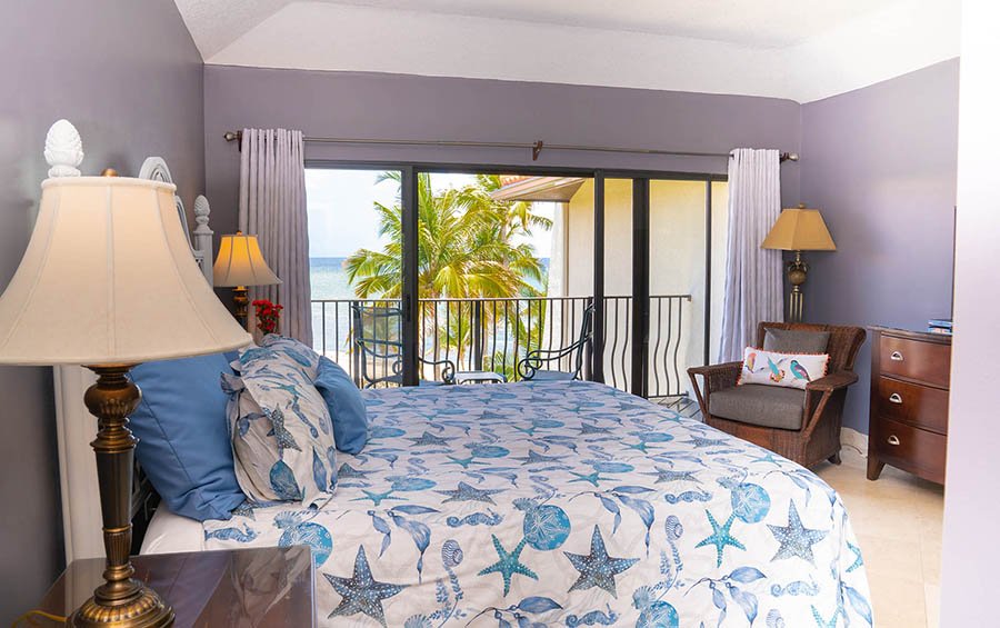 A bedroom with a large bed covered in ocean-themed bedding, including starfish and shells, near a sliding glass door leading to a balcony with a view of palm trees and the ocean.