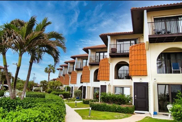 Exterior view of a row of three-story hotel or apartment buildings with balconies, orange-tile accents, and well-maintained landscaping, including palm trees and bushes, under a partly cloudy sky.