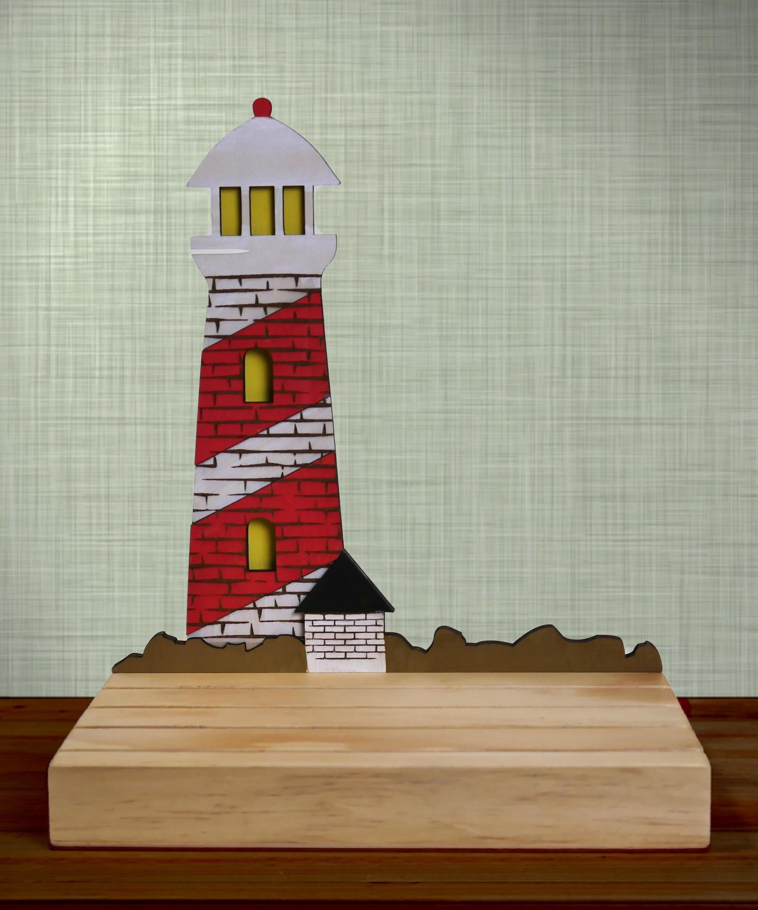 Colorful paper craft of a lighthouse with a red and white striped tower, yellow windows, a small black roof, and a white top, standing on a wooden surface.