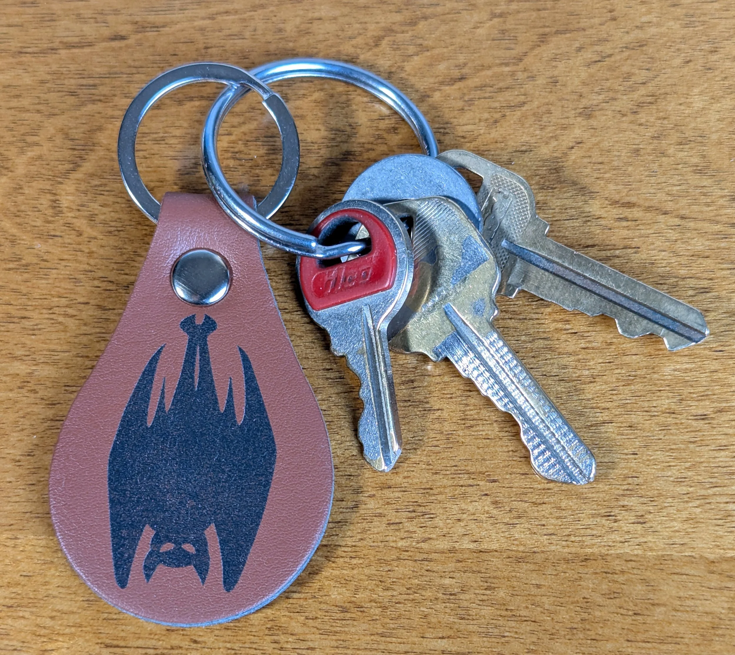 A set of keys on a wooden surface, including a house key, a car key, a key with a red plastic head, and a key with a silver head, attached to a round keyring along with a leather keychain with a logo.