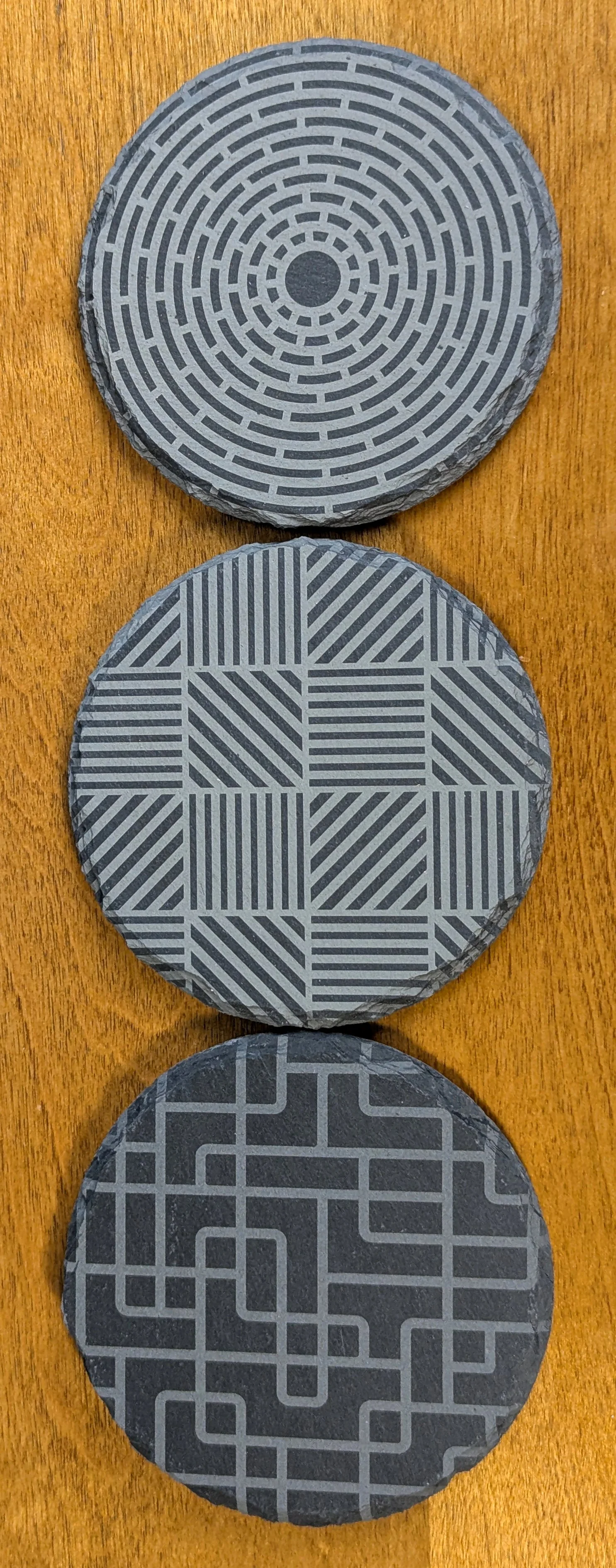 Three round black stones with geometric patterns engraved on their surface, placed on a wooden surface. The patterns include concentric circles, squares with diagonal lines, and interconnected lines.