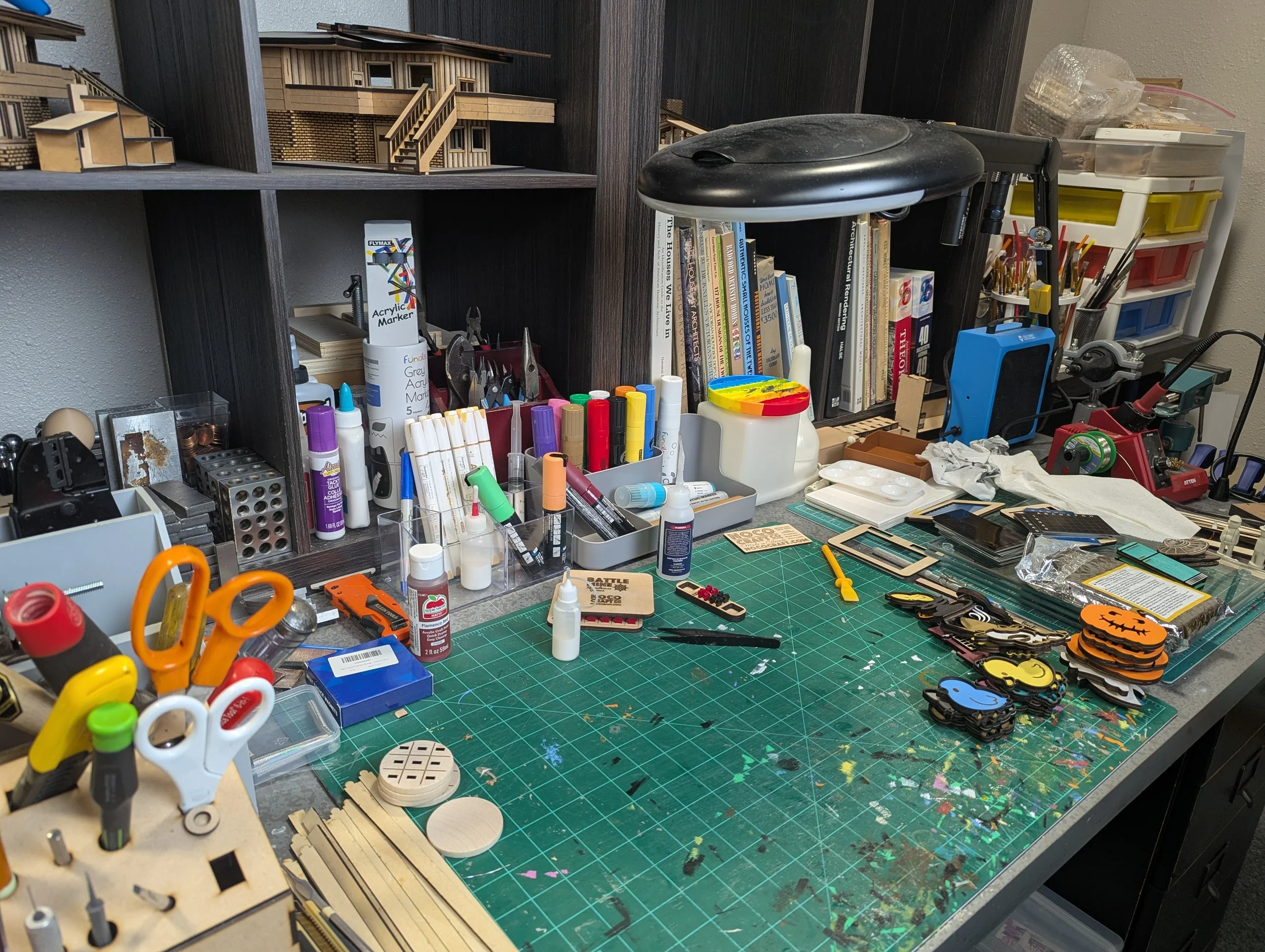 A cluttered worktable with arts and crafts supplies, tools, markers, paint, model pieces, and wooden structures, organized with shelves and storage bins in the background.