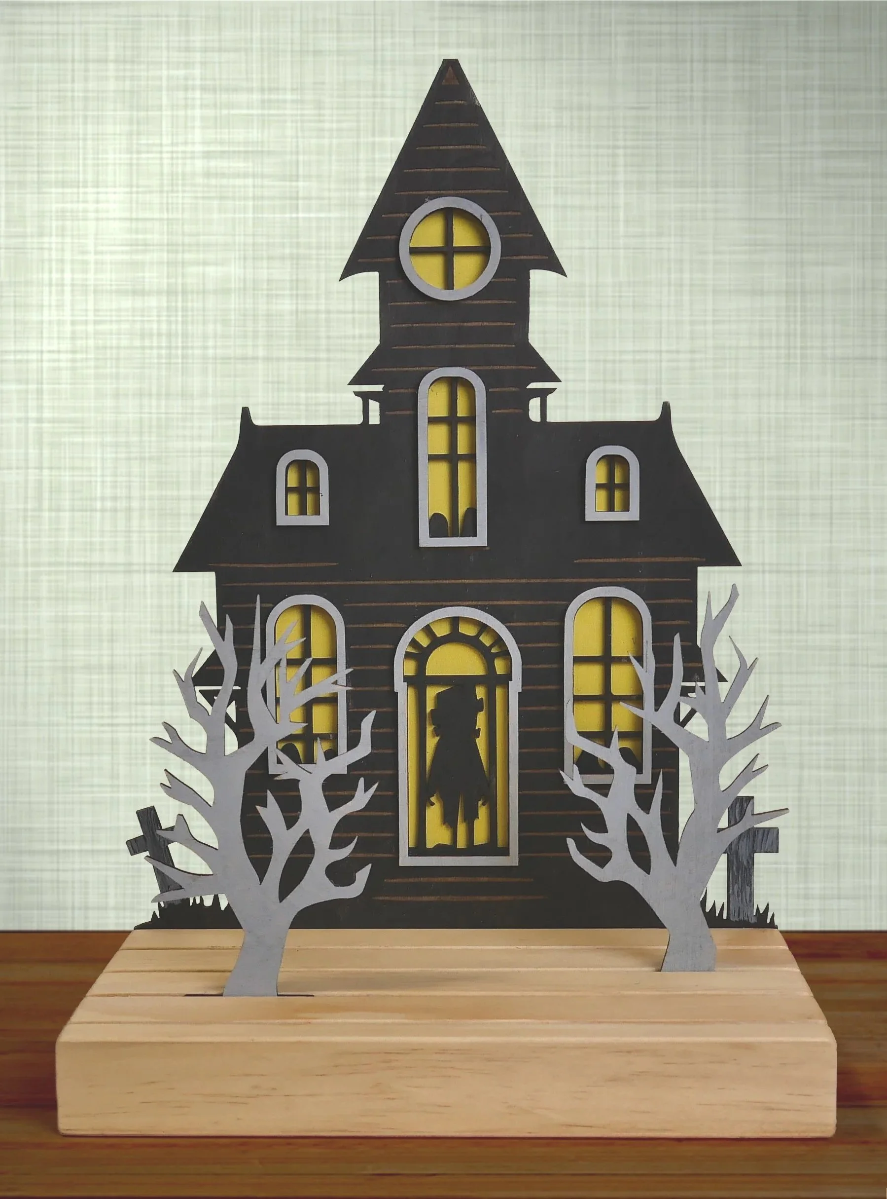A Halloween-themed paper house with dark black walls, yellow windows, and white leafless trees in front, set on a wooden base.