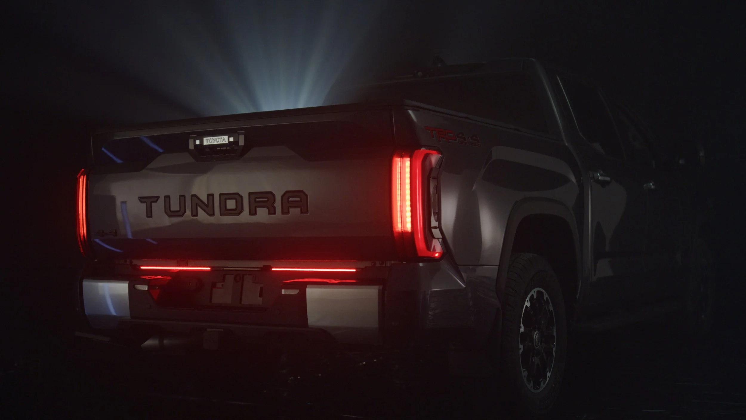 Rear view of a silver Toyota Tundra pickup truck with illuminated tail lights at night, showcasing the Tundra logo on the tailgate and a Toyota badge.