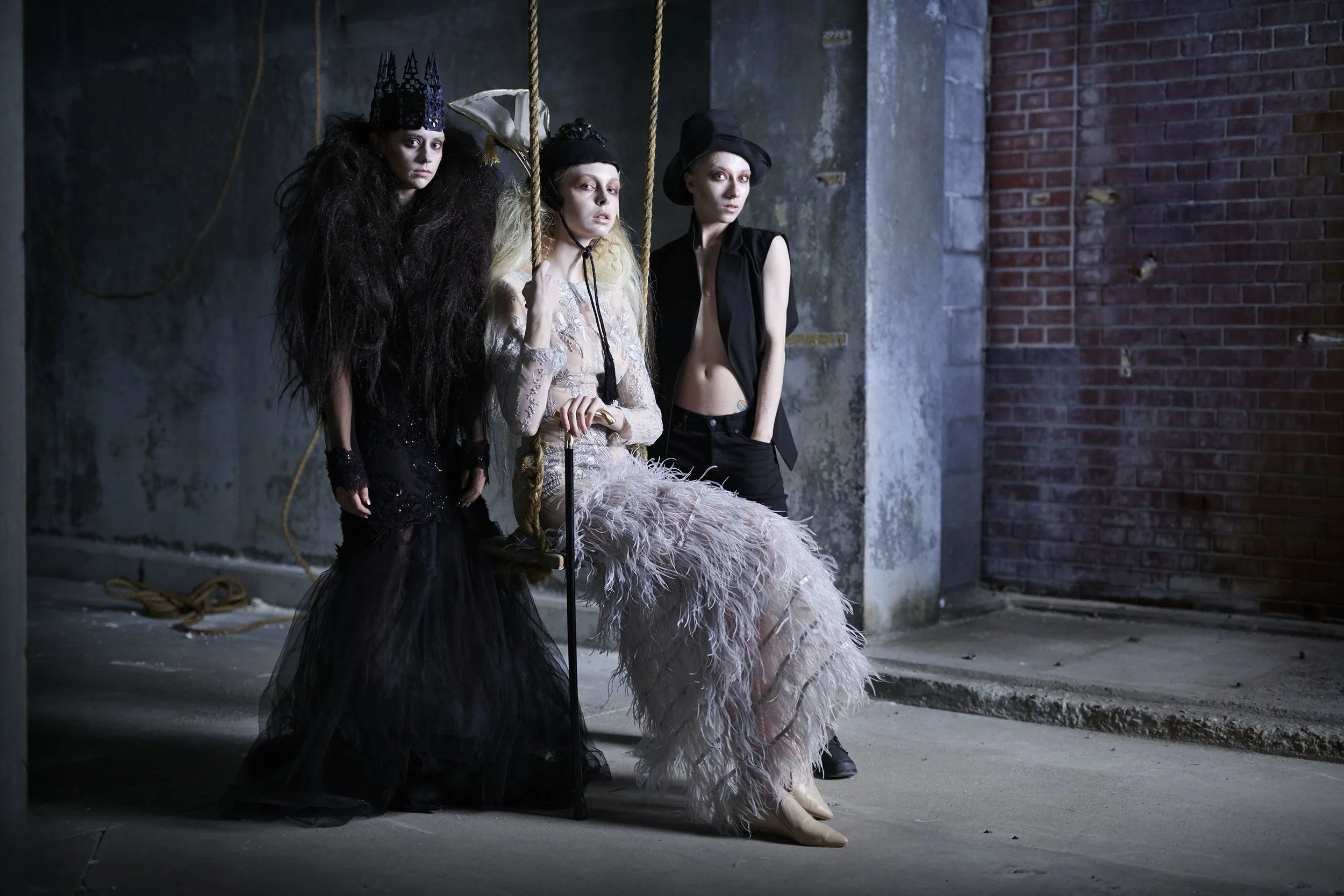 Three women dressed in dark, elaborate costumes, with one wearing a big crown and another in a feathered dress, sitting on a swing in an industrial space with exposed brick and concrete walls.