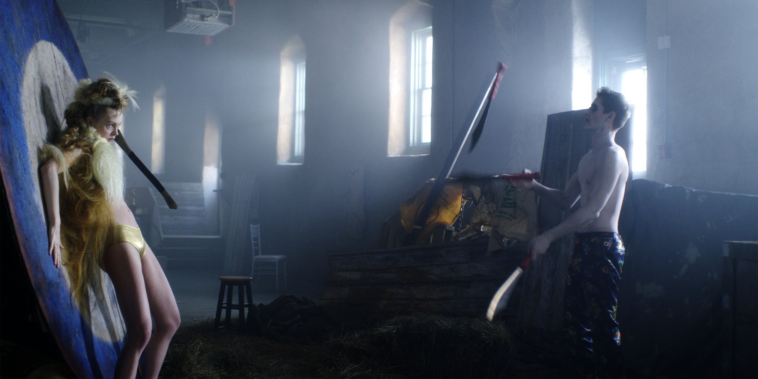 Two young men are in a dimly lit room with sunlight streaming through windows. One young man, shirtless, is standing and holding a shovel. The other young man, with long blonde hair, is leaning against a large, vertical canvas or wall.