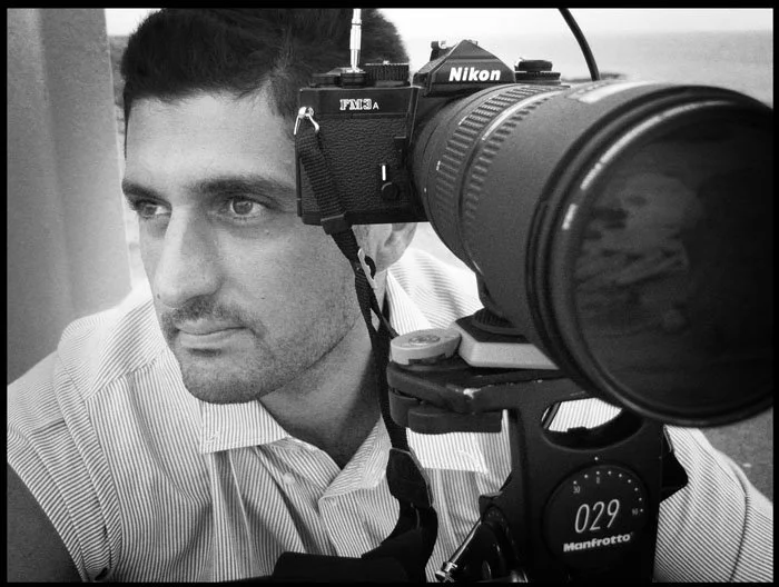 A man with dark hair in a striped shirt looking to the side through the viewfinder of a camera setup for photography.