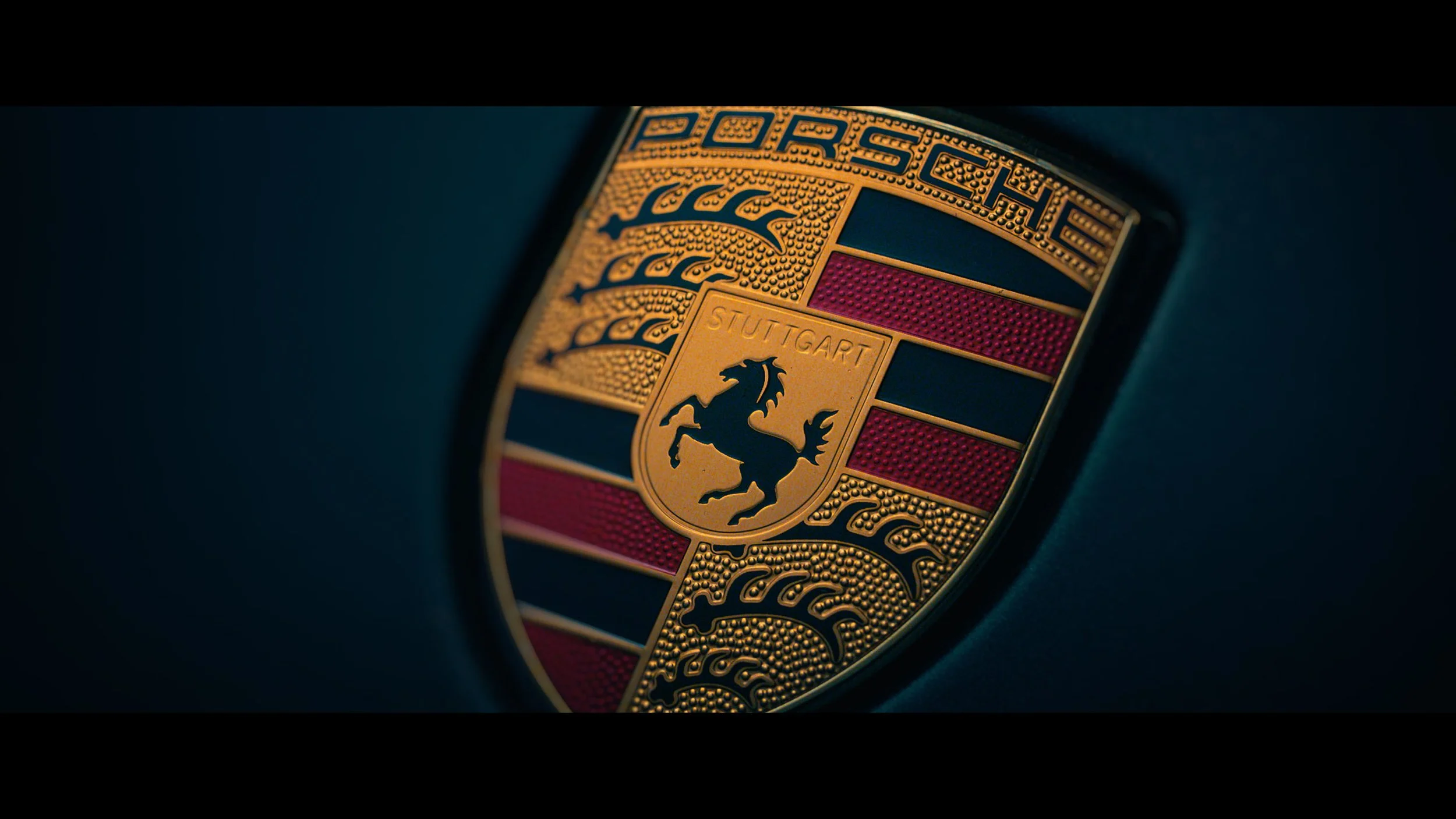 Close-up of a Porsche emblem on a dark background, featuring a black horse within a gold shield, with red and black stripes and the word 'Stuttgart'.
