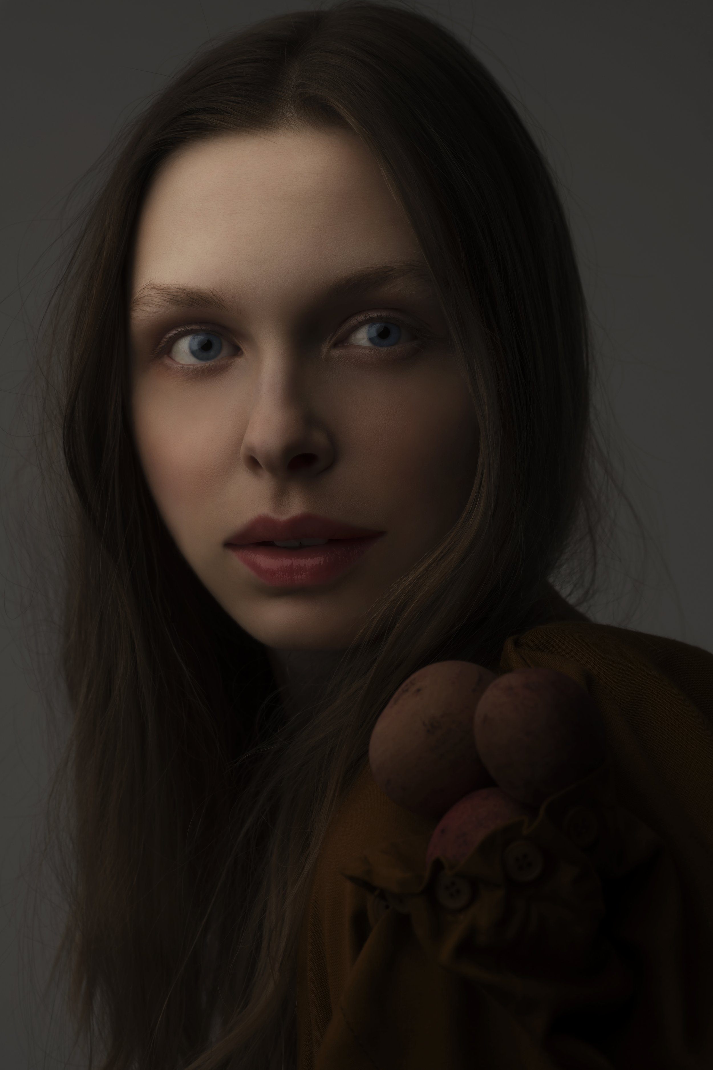 Close-up portrait of a woman with long brown hair and blue eyes, holding grapes, wearing a brown top, with a neutral background.