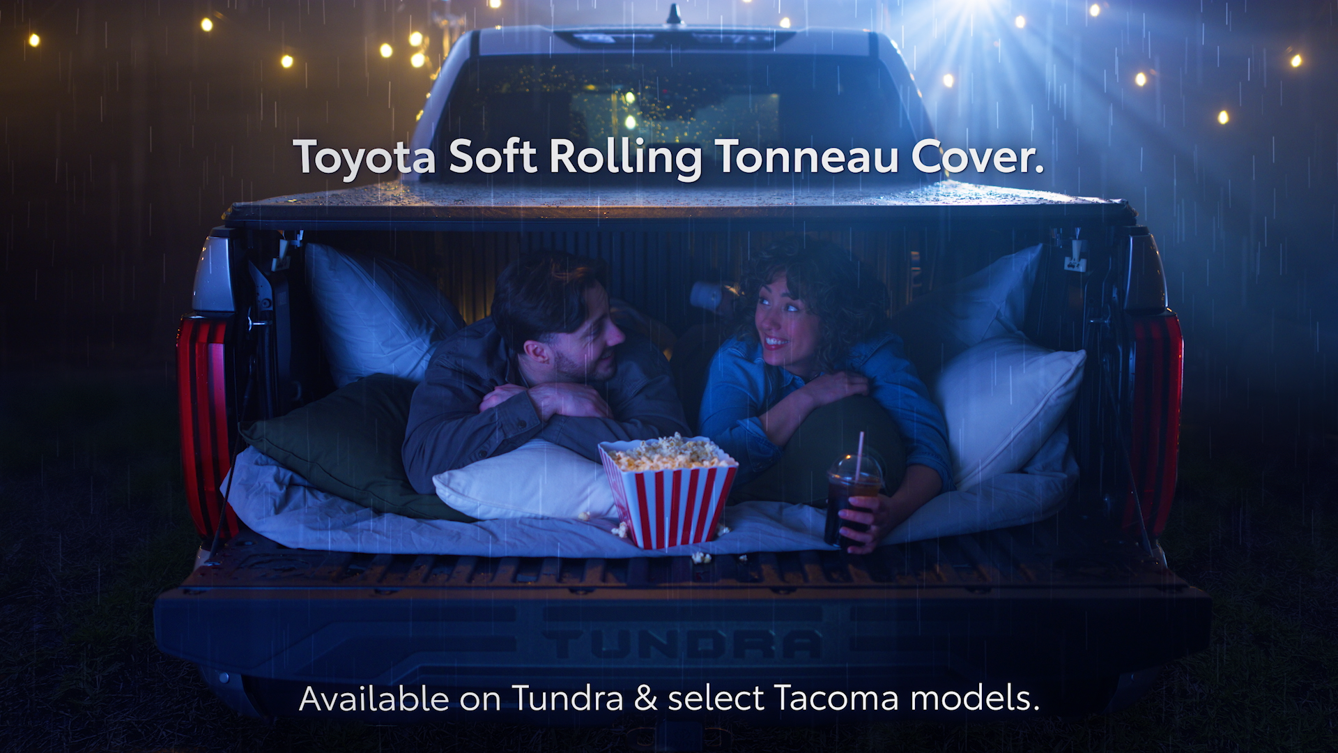 A couple is lying inside the bed of a pickup truck at night in the rain, watching movies. The truck has a soft rolling tonneau cover. They are enjoying popcorn and drinks, with the woman smiling at the man. The scene is illuminated by the truck's interior lights and rain falling around them.
