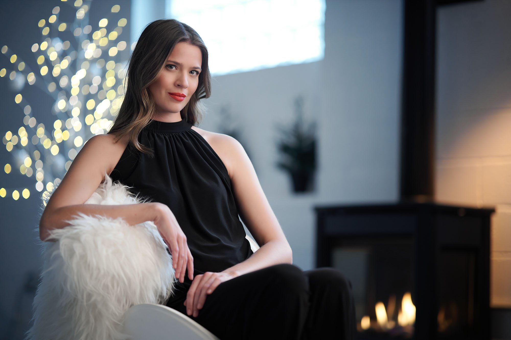 A woman with brown hair, red lipstick, wearing a black sleeveless top, sitting on a white furry chair, with a string of blurred yellow lights and a window in the background.
